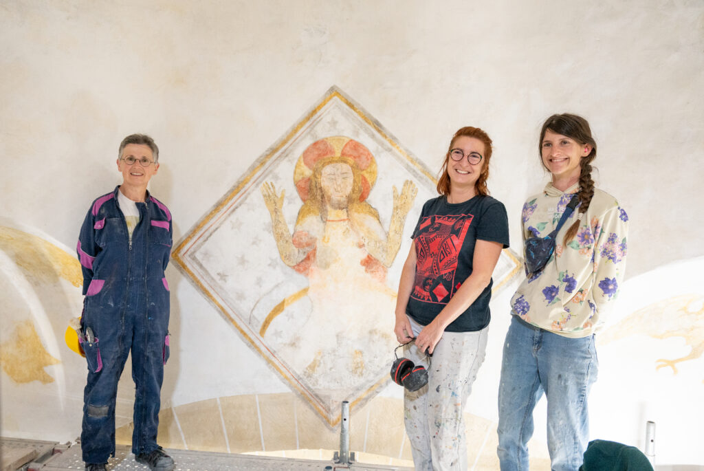 Trois membres de l'équipe de restauration posant devant une peinture murale restaurée dans l'église